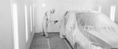 Lackierer in Schutzanzug sprüht einen Abziehlack auf ein abgedecktes Auto in einer professionellen Lackierkabine.